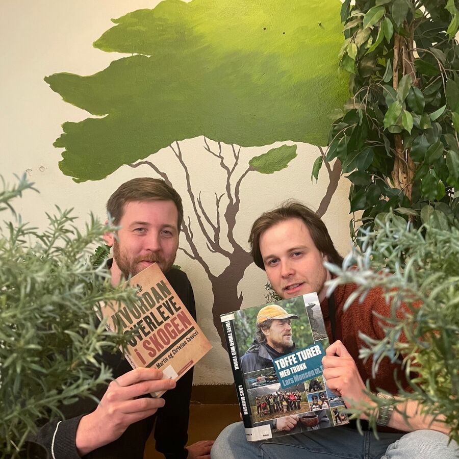 Bilde av Aleksander og Thorbjørn som holder bøkene Hvordan overleve i skogen og tøffe turer med trøkk omgitt av planter og et malt tre på veggen