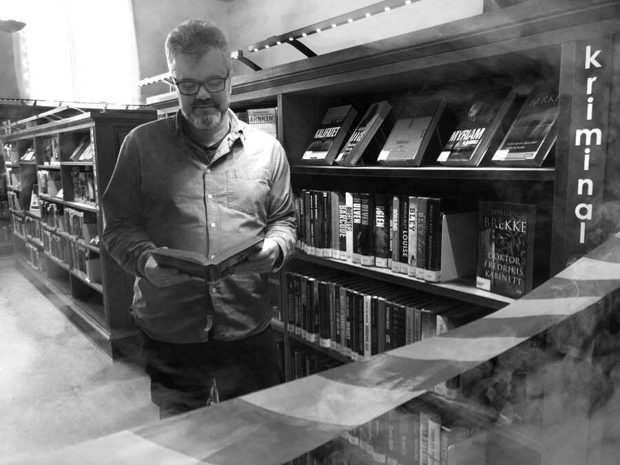 Foto av bibliotekar Dag Einar som leser i en bok mens han står ved siden av hylla med krimbøker bak et sperrebånd og med antydning til røyk i rommet