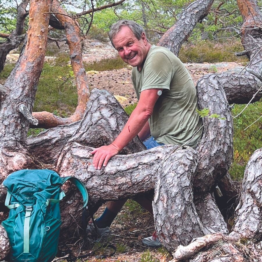 Bilde av Svein Syversen som smiler i kameraet og sitter blant noen lave furugreiner med tursekk