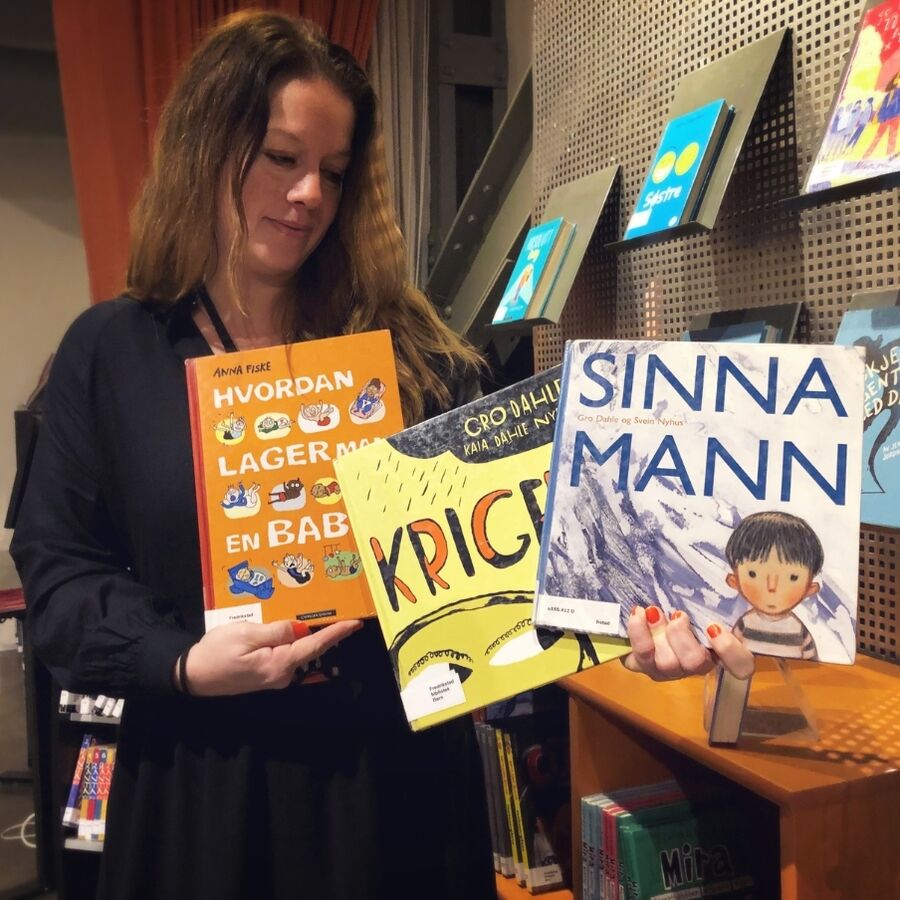 Bilde av bibliotekar Ingvild som holder opp tre barnebøker med titlene Sinna Mann, Krigen og Hvordan lager man en baby?
